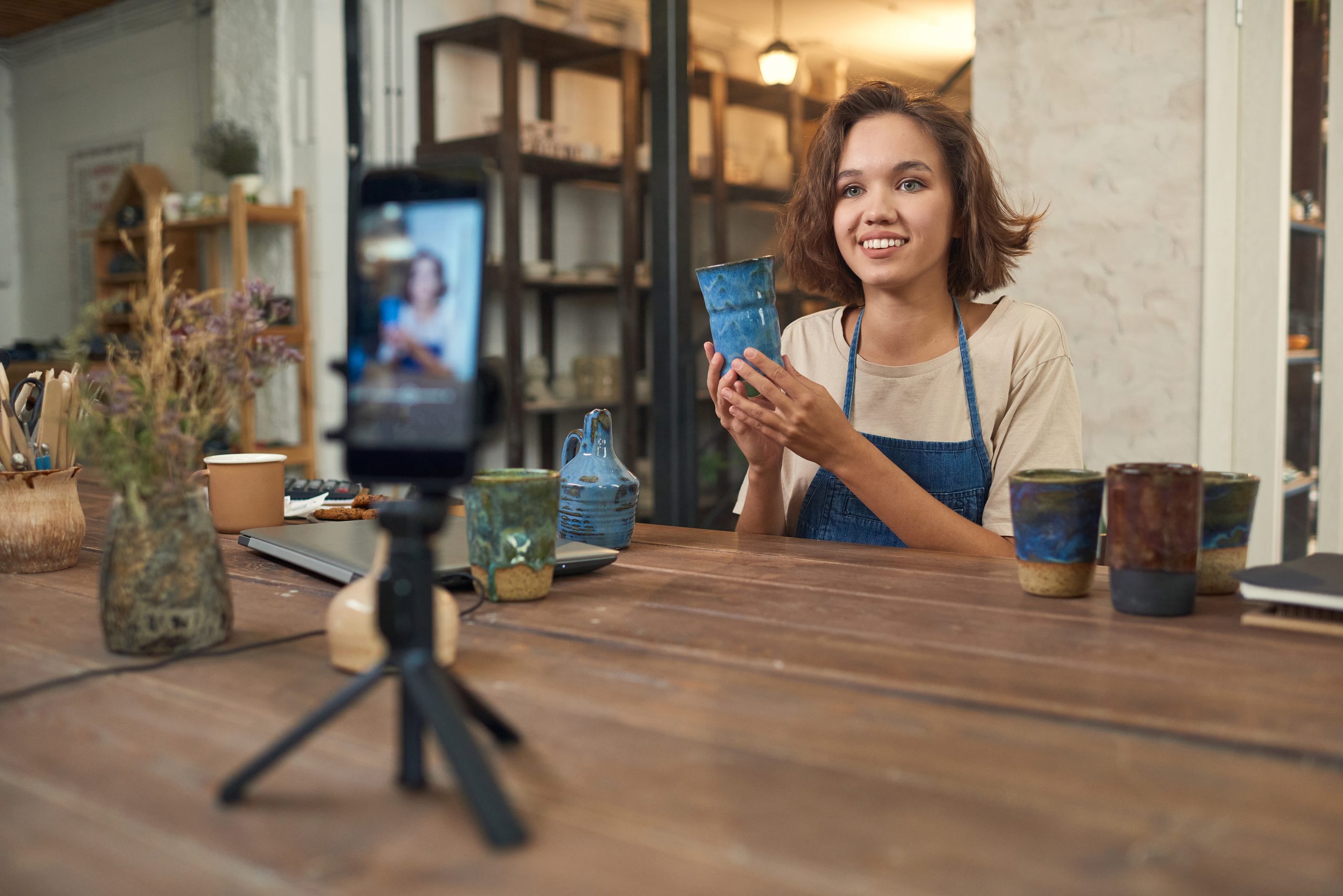 Smiling young Caucasian potter sitting at wooden table in workshop and holding ceramic vase while shooting video for social media on smartphone